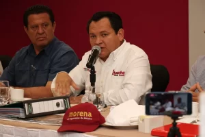 Hombre con camisa blanca hablando en micrófono en reunión con documentos y gorra roja de Morena